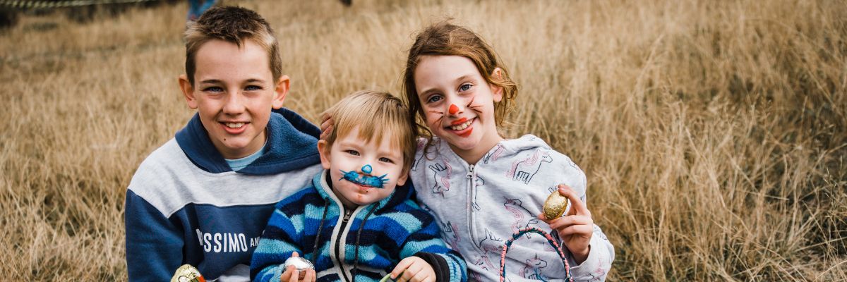 Three children sitting in a field with faces painted as bunnies holding Easter eggs