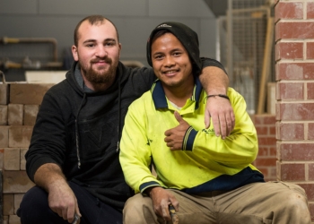 Two people sitting together, one with an arm around the other and one giving the thumbs up