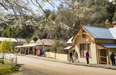 A serene street scene featuring historical wooden buildings under blossoming trees, with people walking along the sidewalk.