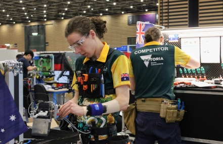 A competitor in a yellow and green uniform labeled  works diligently on machinery at a workbench