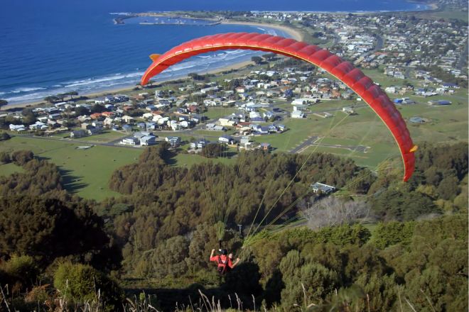 A person paragliding over a coastal town with a clear view of the ocean, residential areas, and green fields.