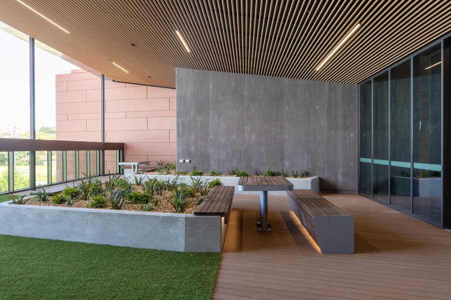 Modern outdoor terrace featuring a wooden deck, raised garden beds with various plants, surrounded by glass walls, under an overhanging ceiling with linear lighting.