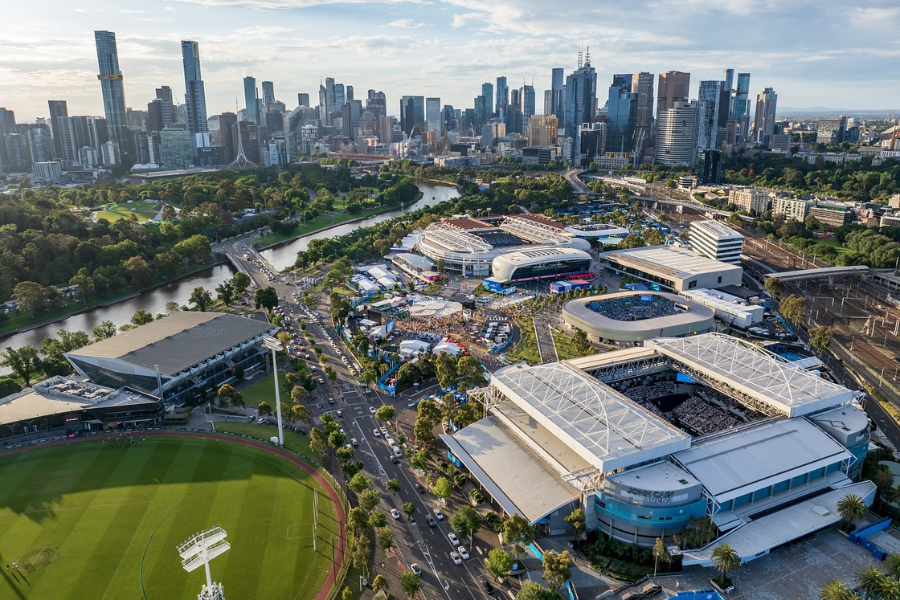 Aerial view of Melbourne Park in Melbourne, Australia, showcasing tennis courts and sports facilities amidst a vibrant city skyline. The Yarra River meanders through the scene, flanked by lush green spaces. Skyscrapers and urban architecture are prominent in the background.