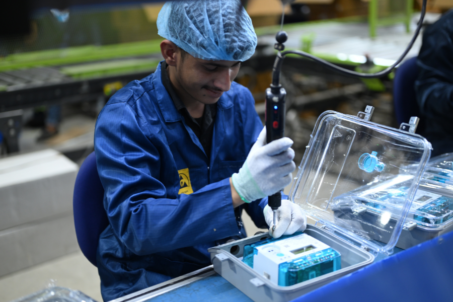 A person in a blue lab coat and hair net works on assembling electronic equipment with a precision tool on a factory production line of Kimbal's smart meters.