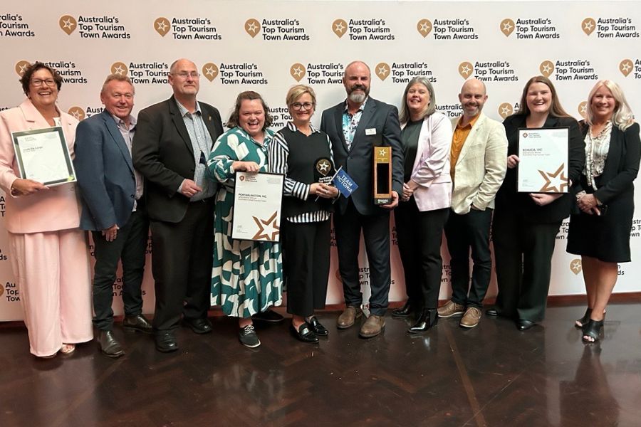 A group of people smiles while holding awards in front of a media wall with the words: Australia's Top Tourism Town Awards