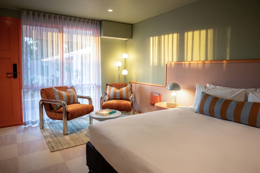 A cozy hotel room with green walls and an orange door, a large bed that has striped pillows, 2 plush orange chairs, a small table with a lamp. Soft natural light filters through sheer curtains covering large windows