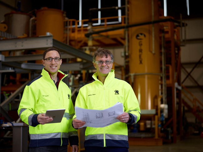 Two workers wearing high visibility jackets stand infront of industrial equipment in a warehouse. They are smiling and looking towards the camera. One is holding a blueprint, the other a computer tablet.