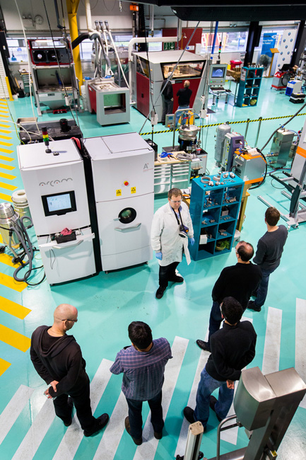 Six people stand in a semicircle on the floor of a manufacturing plant examining the equipment