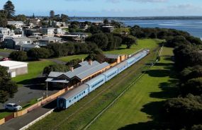 Aerial view of The Bellarine Railway with a train featuring several blue and white carriages. In the background, residential buildings and a calm sea.