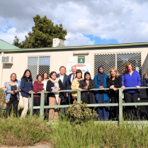 Group of individuals standing in front of a building next to informational signs, smiling for a group photo outdoors.