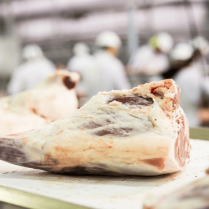 Leg of lamb in foreground with butchers out of focus in background working.