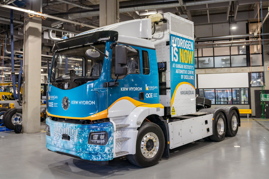A blue and white hydrogen fuel cell truck  showcased in an industrial setting. A banner on the side reads "Hydrogen is now" 