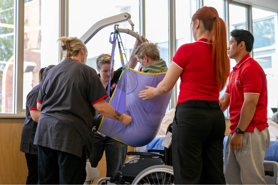 Four healthcare students assist a patient in a sling during a transfer from a wheelchair to a bed.