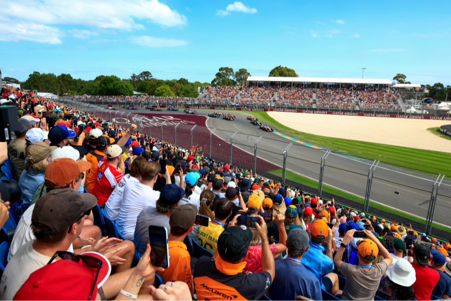 A crowded audience watches a Formula 1 race at Albert Park Circuit in Melbourne. The race cars speed around a bend on the track. Spectators are seated in stands and on a grassy hill, with people capturing the event on their phones.