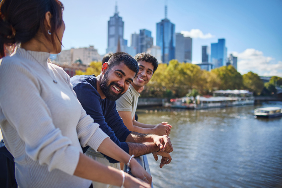 Three international students smiling and leaning on a railing by the Yarra river, with the Melbourne cityscape  in the background. The scene is bright and sunny.