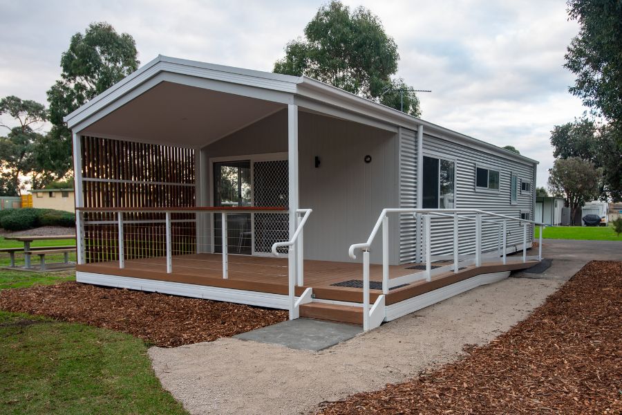 A modern white caravan park cabin with a wooden deck and ramp on landscaped grounds surrounded by trees.