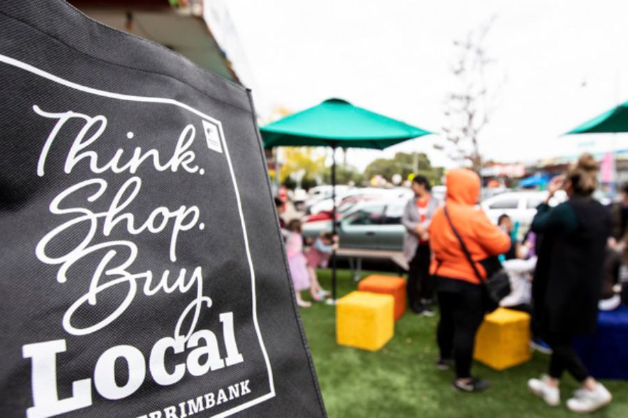 A black reusable shopping bag printed with words: Think. Shop. Buy Local. Brimbank. In the background, people are gathered under green umbrellas, some sitting on coloured blocks underneath. There are parked cars and trees nearby. 
