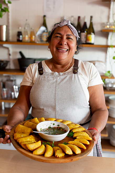 A picture containing a woman smiling, holding a plate of food with shelves in the background