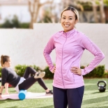 Women in athletic clothing with hands on her hips smiles to camera with gym equipment behind her