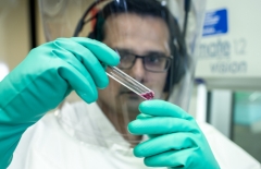 Close up of the gloved hands of a researcher using a pipette. He is wearing a large clear head mask and white gown