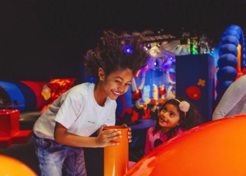 A child with a big smile and wild hair is playing in an indoor playground, leaning over an orange structure.