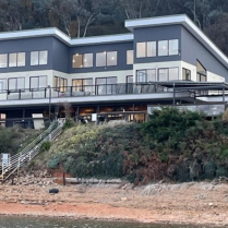 A modern two-story building with large windows and multiple balconies, perched on a sloping hillside by the lakeside