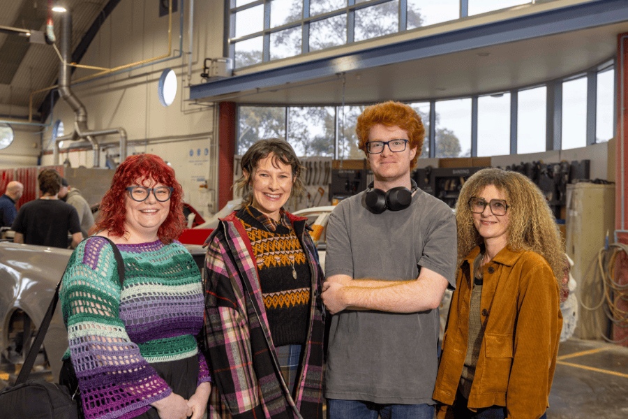 4 students smiling in a workshop environment, with a car and machinery in the background. They are dressed in casual attire, suitable for a workspace.