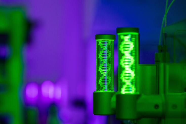 Two glowing green DNA models inside transparent cylinders in a laboratory illuminated with purple and blue lights.