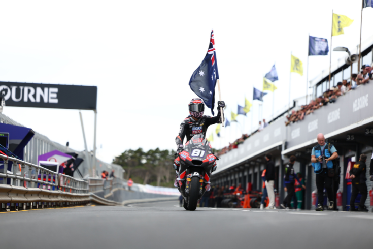 Senna Aguis, motorcyclist, holds an Australian flag while riding on a track, with spectators and Melbourne signage in the background.