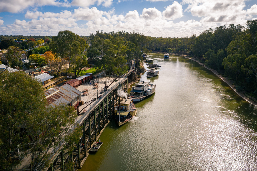 Gold winner of Top Tourism Town - Echuca Aerial view of the Murray River in Echuca, Australia, showing historic paddle steamers docked alongside a wooden wharf. Lush trees line the riverbank under a partly cloudy sky.