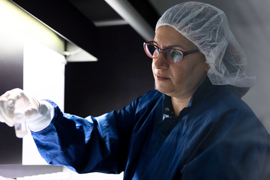 A lab worker in blue scrubs and a disposable hair cover and safety glasses holding a vial at a workstation in a dark lab setting.