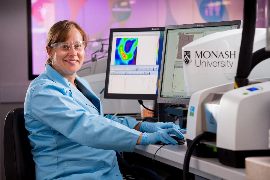 A person is wearing a blue lab coat, gloves and safety glasses sitting at a computer workstation. Monash University equipment is on the table.