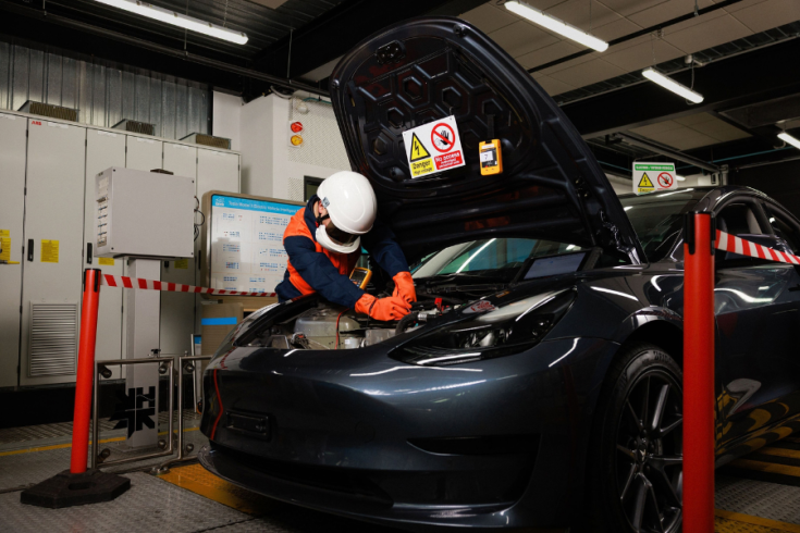 A person wearing safety gear works on the engine of an electric car in an industrial setting. The hood is open, and the area is marked with caution tape and safety signs.