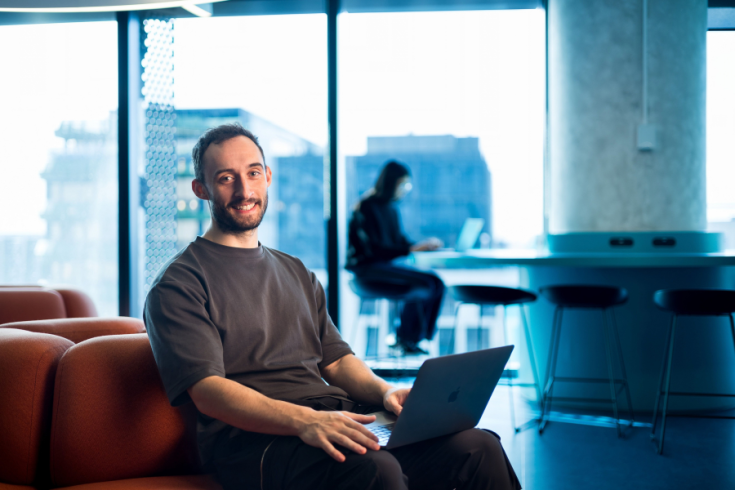 A person smiling while seated on a red couch using a laptop in a bright modern office with large windows, desks, and chairs visible in the background.