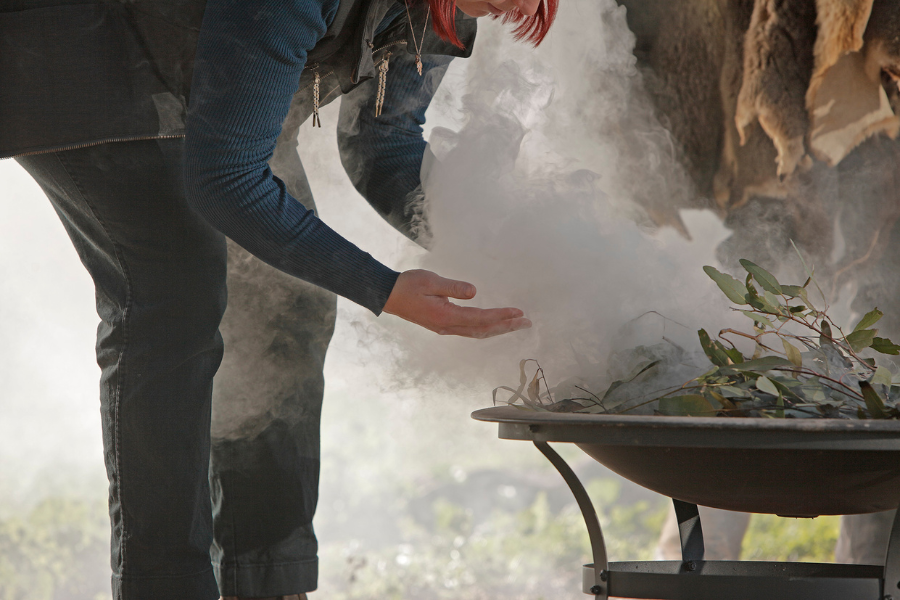 A person is bending over a smoking bowl filled with leaves and twigs, under some hanging animal hides. Sunlight filters through the smoke. A smoking ceremony. 
