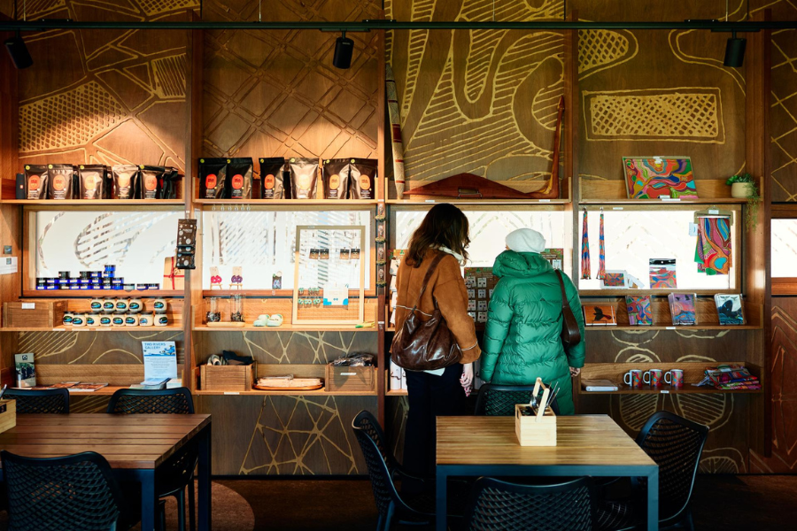 A cozy cafe features intricately carved wooden walls and colourful indigenous artwork. Two people stand near shelves displaying various products like cups and tins.