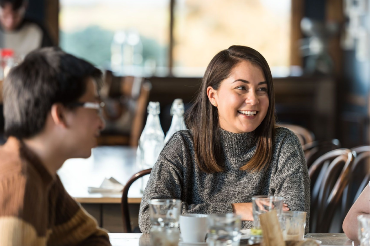Person smiling at a restaurant table while talking with another person, with glassware and bottles on the table and blurred diners in the background.