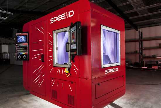 : A worker in a SPEE3D workshop reads a handheld device next to an operating 3D printer.