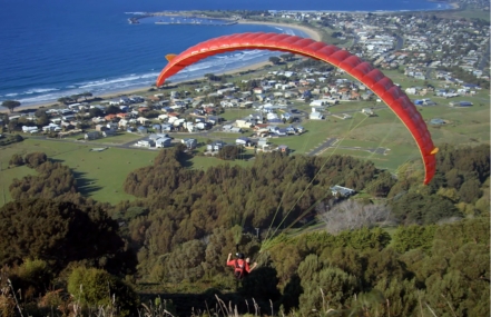 A person paragliding over a coastal town with a clear view of the ocean, residential areas, and green fields.