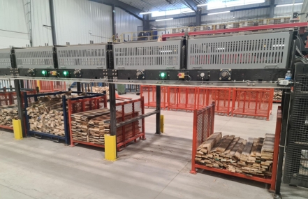 Interior of a warehouse with machinery for processing lumber, featuring stacks of wood on pallets and industrial equipment with green status lights on.