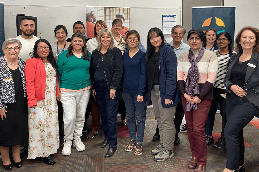 Group of diverse individuals posing for a photo in a classroom setting with a sign that reads "Cultura" in the background.