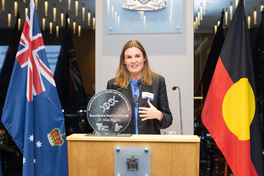 Dr Alisa Higgins at a podium with a microphone, holding an award plaque titled "Research Excellence and Clinical Researcher Award 2024-25". Australian and Aboriginal flags are displayed on either side.