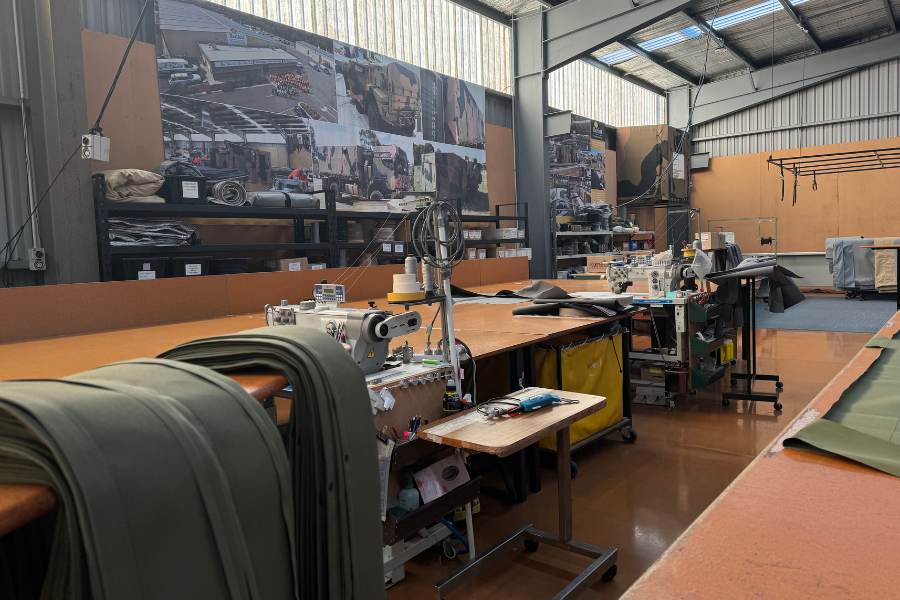Interior of CE Bartlett's busy workshop with fabric rolls, sewing machines, and tables covered with various materials. There are large posters on the wall, depicting industrial scenes.