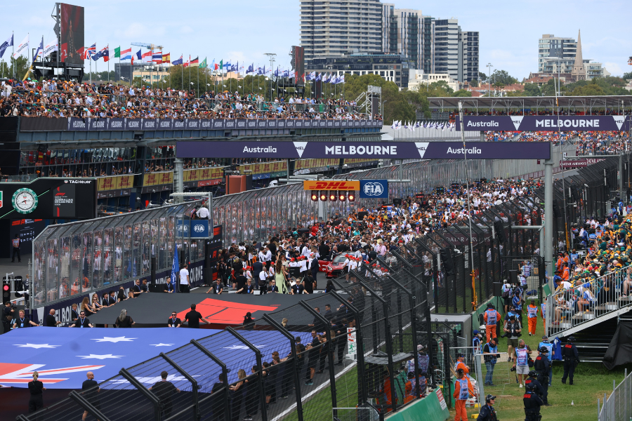 A bustling Formula 1 race event in Melbourne, Australia, with vibrant crowds in the stands. The scene includes a starting grid with cars and attendees preparing under signage for Australia and Melbourne.