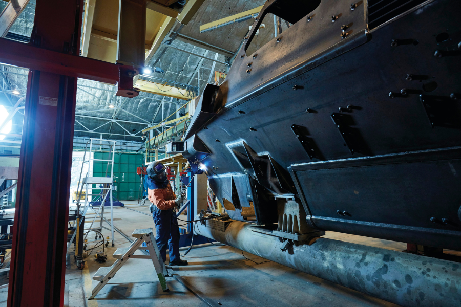 An individual is welding beneath a large industrial vehicle inside a spacious workshop. The area is filled with metal beams, tools, and equipment.
