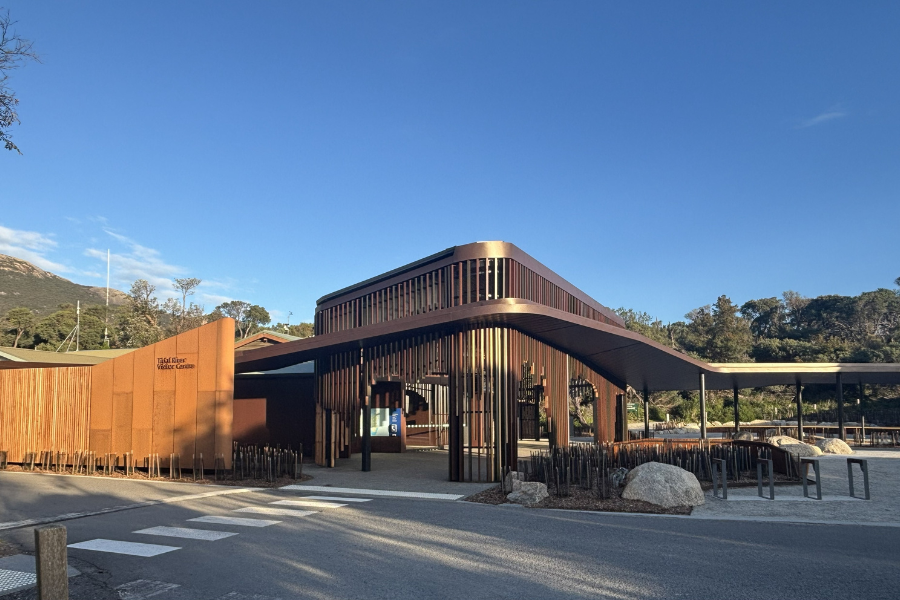 Exterior of the Tidal River Visitor Centre. The building features modern architecture with wood panelling and large glass windows. There is a clear blue sky above, and the surrounding area has some greenery and rocks.