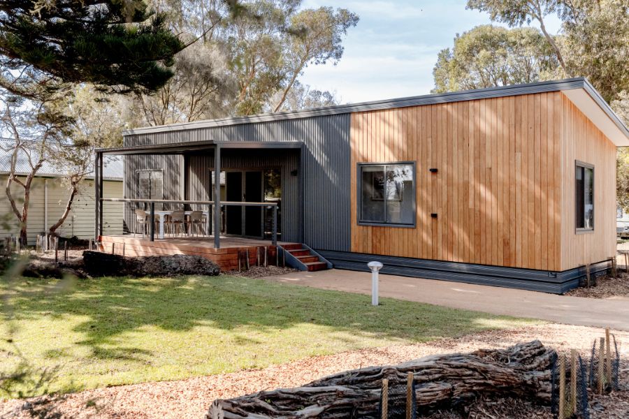 A modern single-story cabin with wood and dark grey siding, featuring a small porch with chairs. The cabin is surrounded by trees and a grass lawn, and a paved walkway leading to the entrance.