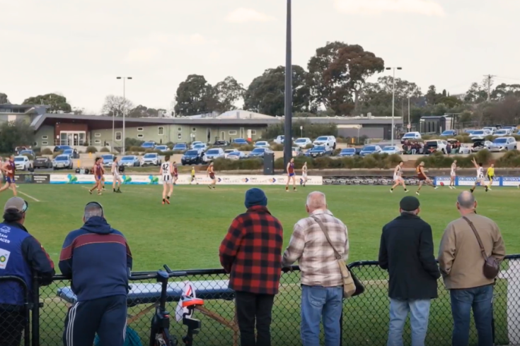 Spectators stand behind a chain-link fence watching a Aussie Rulz football match on a green field, with players in action and teams in blue and red near the Ted Summerton Reserve clubhouse and parking area.