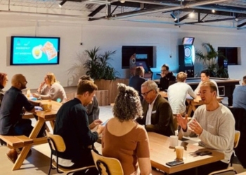 A group of people are gathering in a co-working space, participating in a networking event. There are television screens on the walls, as well as indoor plants in the room. 