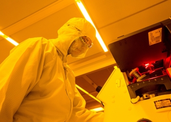 Under orange lights, a person in full medical PPE looks into a microscope